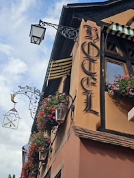 A vintage hotel facade features ornate wrought iron light fixtures and hanging flower baskets. The sign reads 'Hotel' in Gothic lettering, and striped awnings add to the charming, old-world aesthetic.