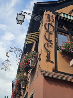 A vintage hotel facade features ornate wrought iron light fixtures and hanging flower baskets. The sign reads 'Hotel' in Gothic lettering, and striped awnings add to the charming, old-world aesthetic.