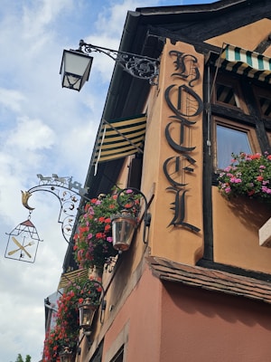 A vintage hotel facade features ornate wrought iron light fixtures and hanging flower baskets. The sign reads 'Hotel' in Gothic lettering, and striped awnings add to the charming, old-world aesthetic.