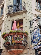 A charming, decorative balcony adorned with potted flowers, including colorful blooms, and a pink umbrella. The building features stone fa&ccedil;ade with detailed iron railings. A vintage sign reads 'Le Petit Schlossberg' with the word 'Caveau' below, attached to the wall next to a classic street lamp.