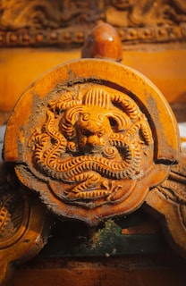 An intricately carved stone or wooden circular emblem featuring a mythical creature, possibly a dragon, with detailed features and swirling patterns. The surface shows signs of age and weathering, giving it a rustic appearance.