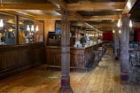 A spacious, warmly-lit pub interior featuring a long wooden bar with high stools. The bar area is adorned with mirrors and elegant light fixtures. Red curtains hang in the background, and wooden beams support the ceiling. The floor is made of wooden planks, enhancing the rustic charm.
