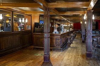 A spacious, warmly-lit pub interior featuring a long wooden bar with high stools. The bar area is adorned with mirrors and elegant light fixtures. Red curtains hang in the background, and wooden beams support the ceiling. The floor is made of wooden planks, enhancing the rustic charm.