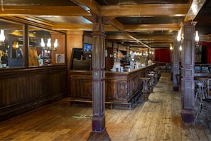 A spacious, warmly-lit pub interior featuring a long wooden bar with high stools. The bar area is adorned with mirrors and elegant light fixtures. Red curtains hang in the background, and wooden beams support the ceiling. The floor is made of wooden planks, enhancing the rustic charm.