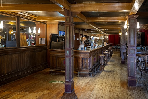 A spacious, warmly-lit pub interior featuring a long wooden bar with high stools. The bar area is adorned with mirrors and elegant light fixtures. Red curtains hang in the background, and wooden beams support the ceiling. The floor is made of wooden planks, enhancing the rustic charm.
