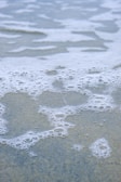 Foamy bubbles floating gently on shallow water create a soft, textured appearance. The water has a translucent quality, and the bubbles form intricate patterns as they spread across the surface.