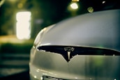 A close-up view of the front of a car featuring the Tesla logo, with a blurred background illuminated by soft, warm lighting.