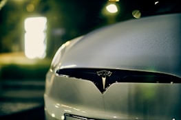 A close-up view of the front of a car featuring the Tesla logo, with a blurred background illuminated by soft, warm lighting.