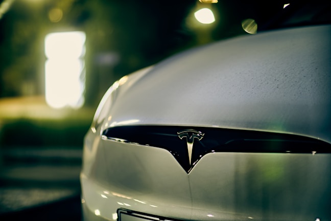 A close-up view of the front of a car featuring the Tesla logo, with a blurred background illuminated by soft, warm lighting.