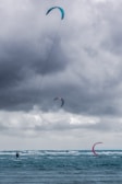 A group of kite surfers are gliding across rough seas, riding the waves under a cloudy, overcast sky. The kites are vibrant in colors, contrasting against the dark clouds, with visible lines connecting the kites to the surfers.