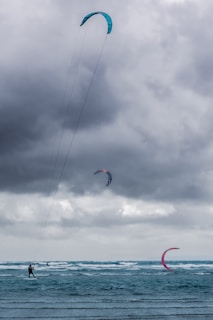 A group of kite surfers are gliding across rough seas, riding the waves under a cloudy, overcast sky. The kites are vibrant in colors, contrasting against the dark clouds, with visible lines connecting the kites to the surfers.