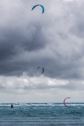 A group of kite surfers are gliding across rough seas, riding the waves under a cloudy, overcast sky. The kites are vibrant in colors, contrasting against the dark clouds, with visible lines connecting the kites to the surfers.