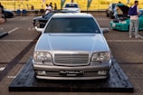 A silver Mercedes-Benz car is prominently displayed on a wooden platform in a parking area. There are other vehicles and a few people in the background, with a person inspecting the engine of a turquoise car.