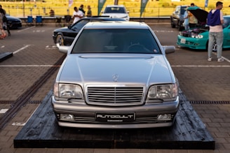 A silver Mercedes-Benz car is prominently displayed on a wooden platform in a parking area. There are other vehicles and a few people in the background, with a person inspecting the engine of a turquoise car.