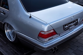 A silver luxury car with a sleek design is parked on a paved surface. The rear view highlights the car's smooth metallic finish, distinctive taillights, dual exhaust pipes, and an emblem on the trunk. The surroundings include cobblestone pavement and a wooden surface nearby.