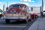 An antique car with a rusted finish is parked outdoors at an event. The vehicle has prominent chrome accents and is surrounded by modern infrastructure, including a building and tents. There are people nearby, and banners or flags are visible around the area.
