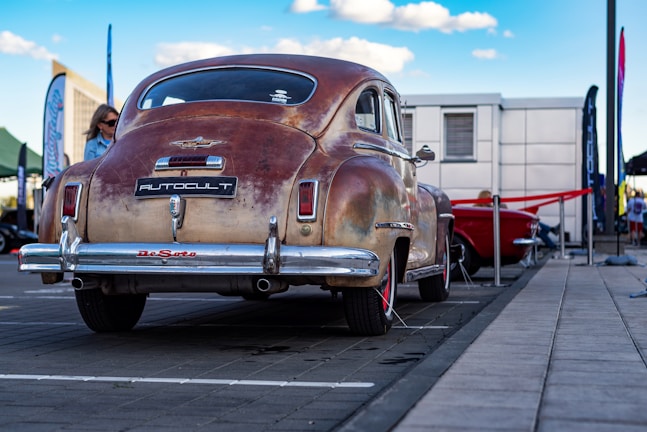 An antique car with a rusted finish is parked outdoors at an event. The vehicle has prominent chrome accents and is surrounded by modern infrastructure, including a building and tents. There are people nearby, and banners or flags are visible around the area.