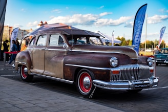A vintage, well-worn classic car with a rusty patina finish and red-rimmed wheels is parked on a paved surface. The background includes flags, a tent, and blurred people, indicating an outdoor event or exhibition. The sky is blue with scattered clouds, and there are buildings and trees visible in the distance.
