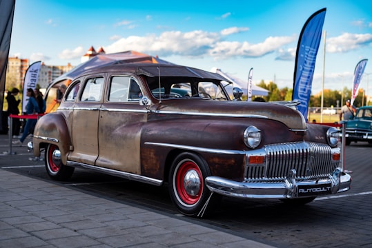 A vintage, well-worn classic car with a rusty patina finish and red-rimmed wheels is parked on a paved surface. The background includes flags, a tent, and blurred people, indicating an outdoor event or exhibition. The sky is blue with scattered clouds, and there are buildings and trees visible in the distance.