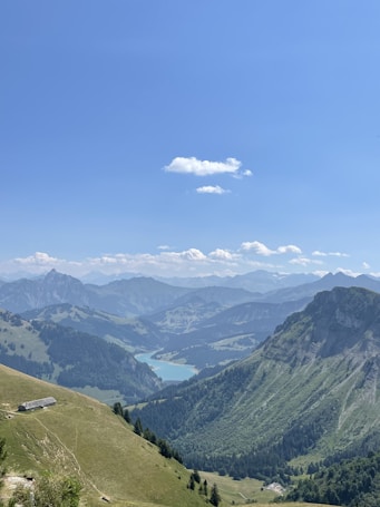 A breathtaking panorama of lush green mountains and valleys with a turquoise lake nestled in the middle. The sky is clear with scattered clouds, providing a serene backdrop to the majestic peaks in the distance. A small structure is visible on a hillside, indicating a remote and peaceful location.