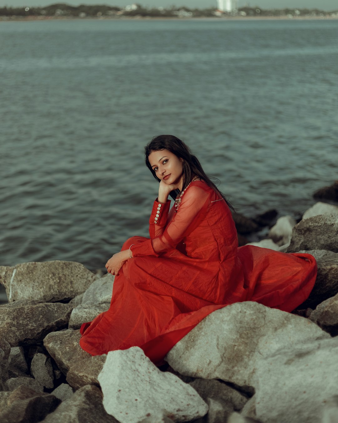 a woman in a red dress sitting on rocks by the water