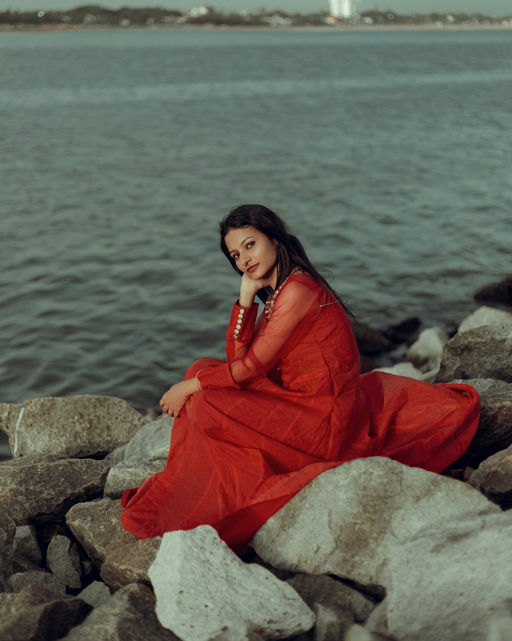 a woman in a red dress sitting on rocks by the water