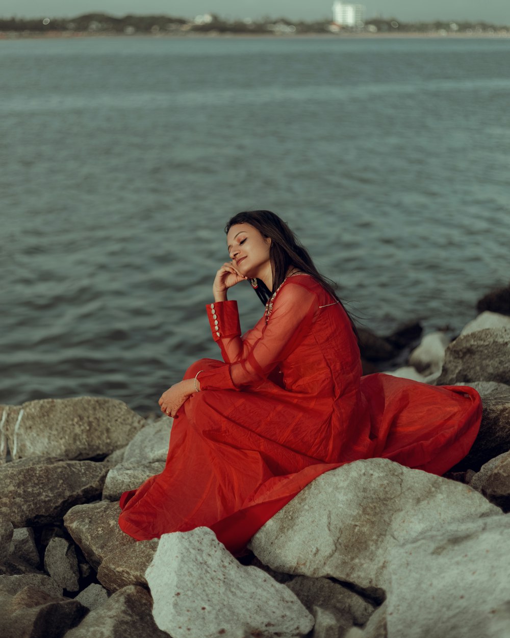 a woman in a red dress sitting on rocks by the water