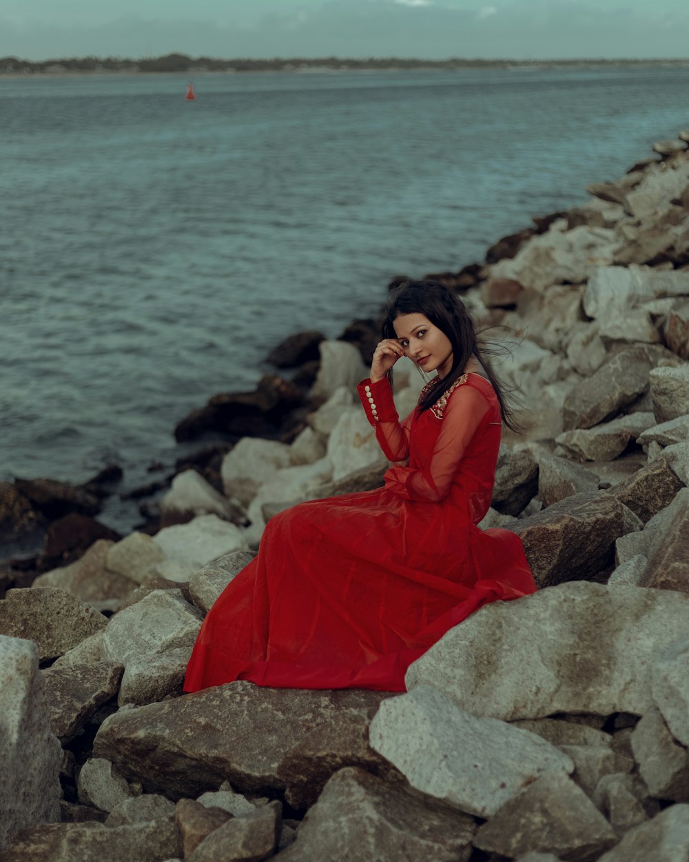 a woman in a red dress sitting on rocks by the water