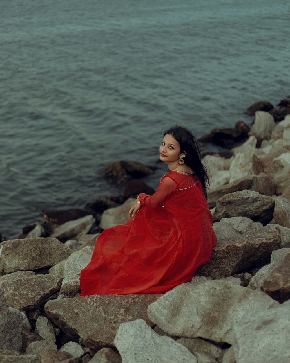 a woman in a red dress sitting on rocks by the water