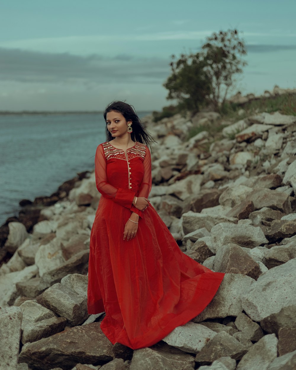 a woman in a red dress standing on rocks by the water