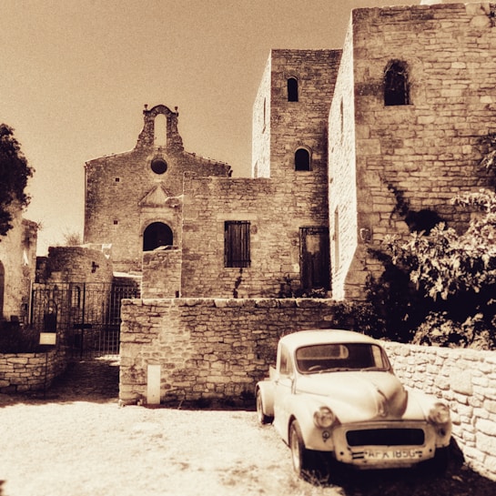 A sepia-toned photograph of old stone buildings, possibly part of a historic village or town. The architecture features arched windows and a small church with a bell tower. In the foreground, there is a vintage car parked near the buildings, enhancing the nostalgic atmosphere.
