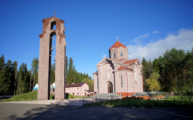 A large stone church with a prominent steeple and red-tiled roof sits in front of a grove of green fir trees under a clear blue sky. Adjacent to the church is a tall, open bell tower structure made of stone, housing a single bell. A small pink building with a brown roof is visible to the left, partially covered by trees.