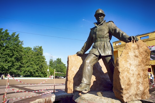 A bronze statue of a miner stands in a dynamic pose, leaning slightly forward with one leg on a large stone. The figure is depicted wearing a helmet and work attire, set against a backdrop of blue sky and lush green trees. Two large boulders are positioned on either side of the statue, and the area is partially cordoned off with a red and white tape.