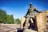 A bronze statue of a miner stands in a dynamic pose, leaning slightly forward with one leg on a large stone. The figure is depicted wearing a helmet and work attire, set against a backdrop of blue sky and lush green trees. Two large boulders are positioned on either side of the statue, and the area is partially cordoned off with a red and white tape.