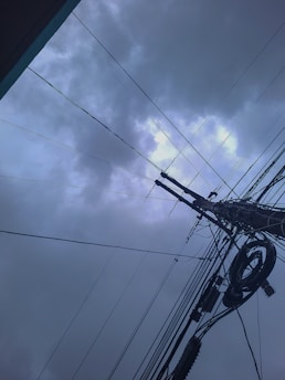 A network of overhead power lines crisscrosses against a backdrop of overcast sky filled with dark, thick clouds. The cables intersect at various angles, converging at a utility pole. The image evokes a sense of industrial complexity against a moody sky.