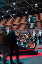 A motorcycle displayed on a platform surrounded by people at an indoor exhibition. Overhead signage indicates various activities such as motocross and roadshows. The area is well-lit with ceiling lights and features modern architecture.