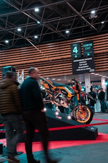 A motorcycle displayed on a platform surrounded by people at an indoor exhibition. Overhead signage indicates various activities such as motocross and roadshows. The area is well-lit with ceiling lights and features modern architecture.