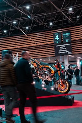 A motorcycle displayed on a platform surrounded by people at an indoor exhibition. Overhead signage indicates various activities such as motocross and roadshows. The area is well-lit with ceiling lights and features modern architecture.
