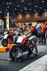 A person is sitting on an orange and black motorcycle displayed indoors at an exhibition or showroom. Several other people are seen interacting and observing various motorcycles in the background. The environment is well-lit with overhead lighting, and the space is designed with modern aesthetics and informational displays.