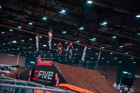 Several motorcyclists are airborne, performing stunts indoors at a motocross event. The venue is equipped with artificial lights and banners, and the ramp is made of dirt. A crowd observes from behind barriers.