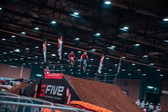 Several motorcyclists are airborne, performing stunts indoors at a motocross event. The venue is equipped with artificial lights and banners, and the ramp is made of dirt. A crowd observes from behind barriers.