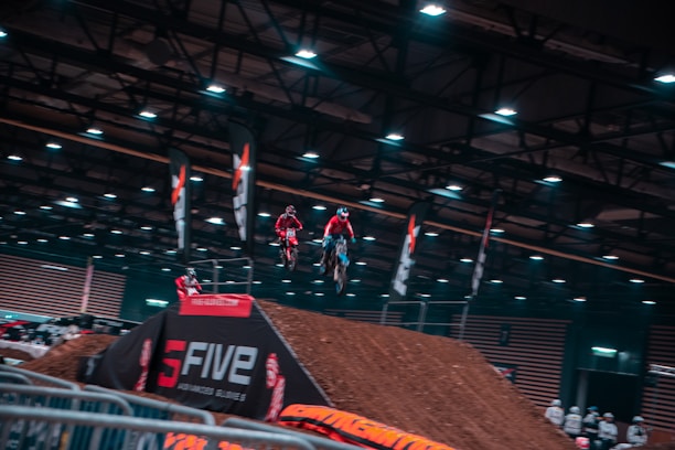 Several motorcyclists are airborne, performing stunts indoors at a motocross event. The venue is equipped with artificial lights and banners, and the ramp is made of dirt. A crowd observes from behind barriers.