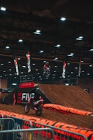 A motocross event indoors, featuring dirt ramps and barriers. Two motorcyclists wearing helmets and gear are actively participating, one is airborne performing a jump while another rides down a ramp. The area is lit with overhead lights and lined with branding banners and caution tape.
