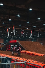 A motocross event indoors, featuring dirt ramps and barriers. Two motorcyclists wearing helmets and gear are actively participating, one is airborne performing a jump while another rides down a ramp. The area is lit with overhead lights and lined with branding banners and caution tape.