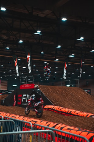A motocross event indoors, featuring dirt ramps and barriers. Two motorcyclists wearing helmets and gear are actively participating, one is airborne performing a jump while another rides down a ramp. The area is lit with overhead lights and lined with branding banners and caution tape.