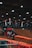 A motocross event indoors, featuring dirt ramps and barriers. Two motorcyclists wearing helmets and gear are actively participating, one is airborne performing a jump while another rides down a ramp. The area is lit with overhead lights and lined with branding banners and caution tape.