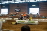 A motorcycle race taking place indoors on a dirt track, with several riders in motion. The setting includes barriers and ramps, and there is a vibrant atmosphere with spectators in the background. The riders are wearing helmets and racing gear.