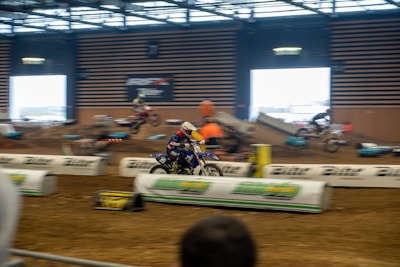 A motorcycle race taking place indoors on a dirt track, with several riders in motion. The setting includes barriers and ramps, and there is a vibrant atmosphere with spectators in the background. The riders are wearing helmets and racing gear.
