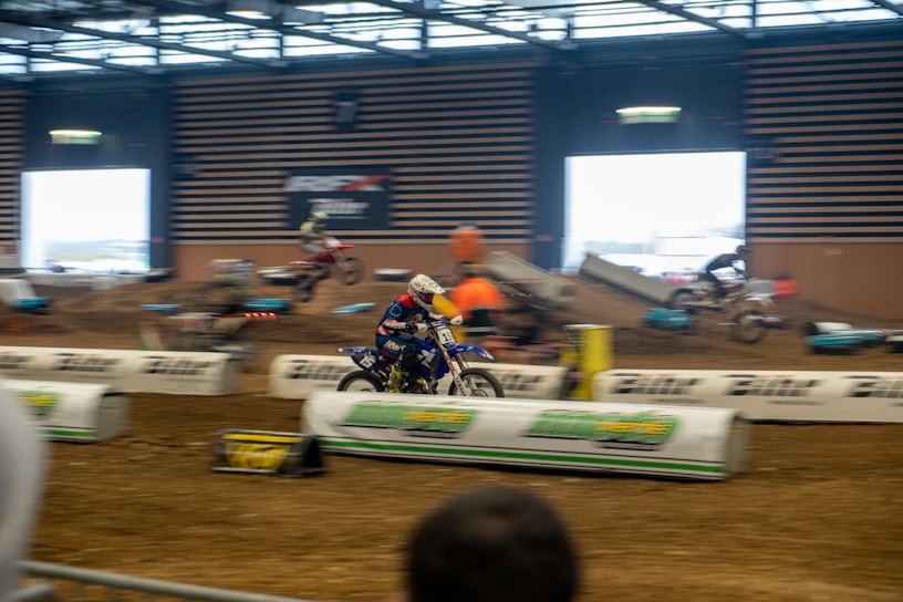 A motorcycle race taking place indoors on a dirt track, with several riders in motion. The setting includes barriers and ramps, and there is a vibrant atmosphere with spectators in the background. The riders are wearing helmets and racing gear.
