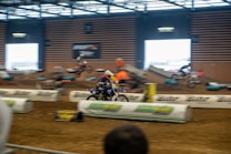 A motorcycle race taking place indoors on a dirt track, with several riders in motion. The setting includes barriers and ramps, and there is a vibrant atmosphere with spectators in the background. The riders are wearing helmets and racing gear.